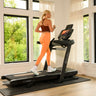 A woman in athletic wear runs on a BowFlex T16 Treadmill in a bright room with large windows and green outdoor views.