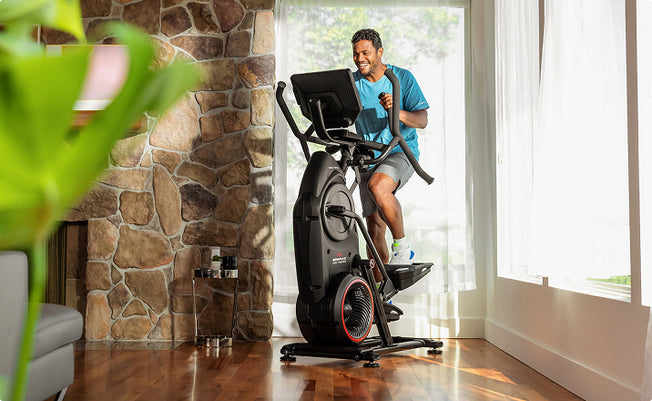A man in athletic wear works out on a BowFlex Max Total 16 in a bright room with large windows.