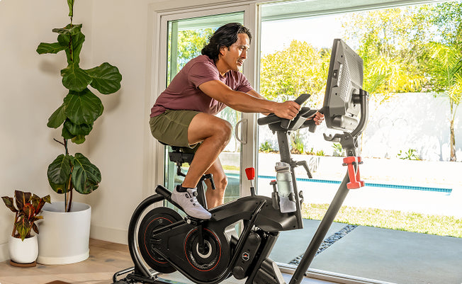 Man using the BowFlex VeloCore Bike 22 in a home setting, with a pool in a yard visible through large windows behind him.