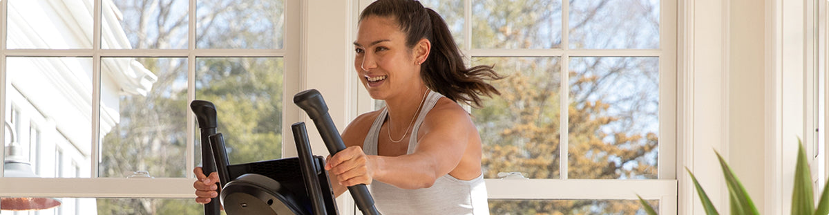 A woman is exercising on a BowFlex Max Trainer M6 in a bright room with large windows.