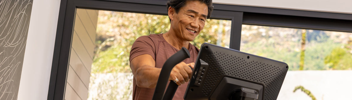 Man smiling while exercising on the BowFlex Max Total 16 in a home setting with a blurred outdoor background visible through the windows.