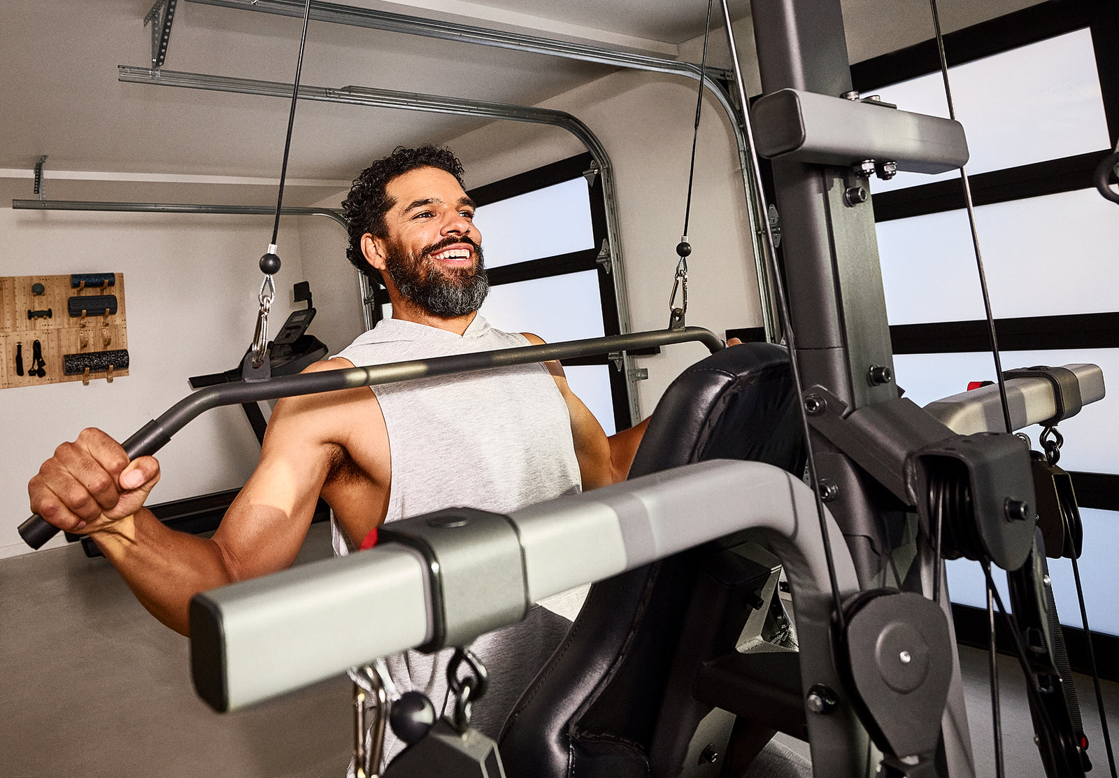 A man with a beard and a gray tank top using the steel curved lat bar on a BowFlex Xtreme 2 SE Home Gym in a garage with garage door windows.