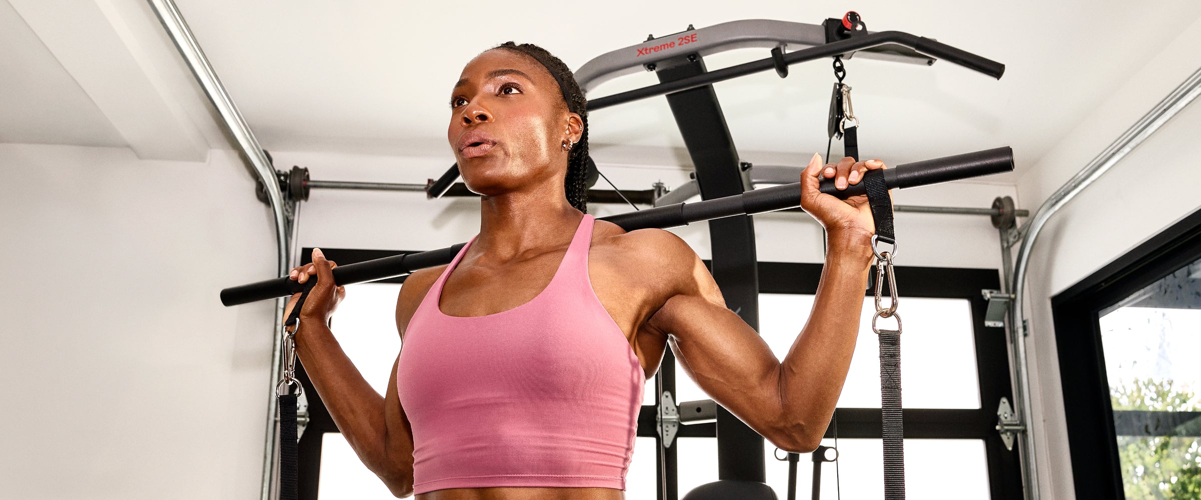 A woman in a pink sports tank top using the 6-grip steel straight bar attachment on the BowFlex Xtreme 2 SE Home Gym.