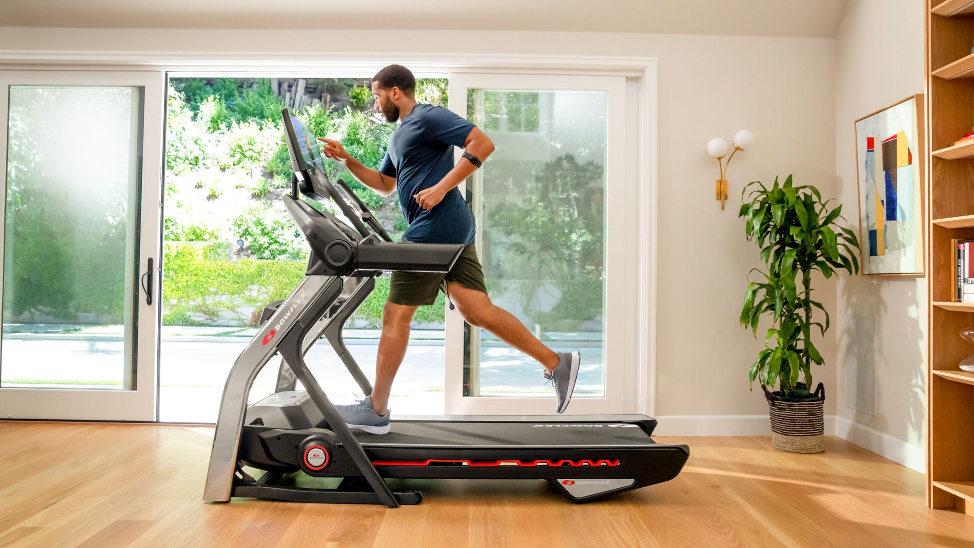 Man running on a treadmill in a home setting with large windows and indoor plants.